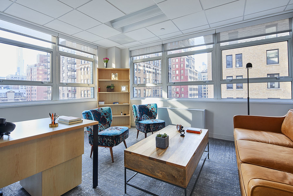 image005 A bright, modern therapy office at The Motherhood Center. The room features large windows with an urban cityscape view, a wooden desk, cozy chairs with floral upholstery, and a leather sofa. The space is inviting and designed to create a comfortable and calming environment for therapy sessions.