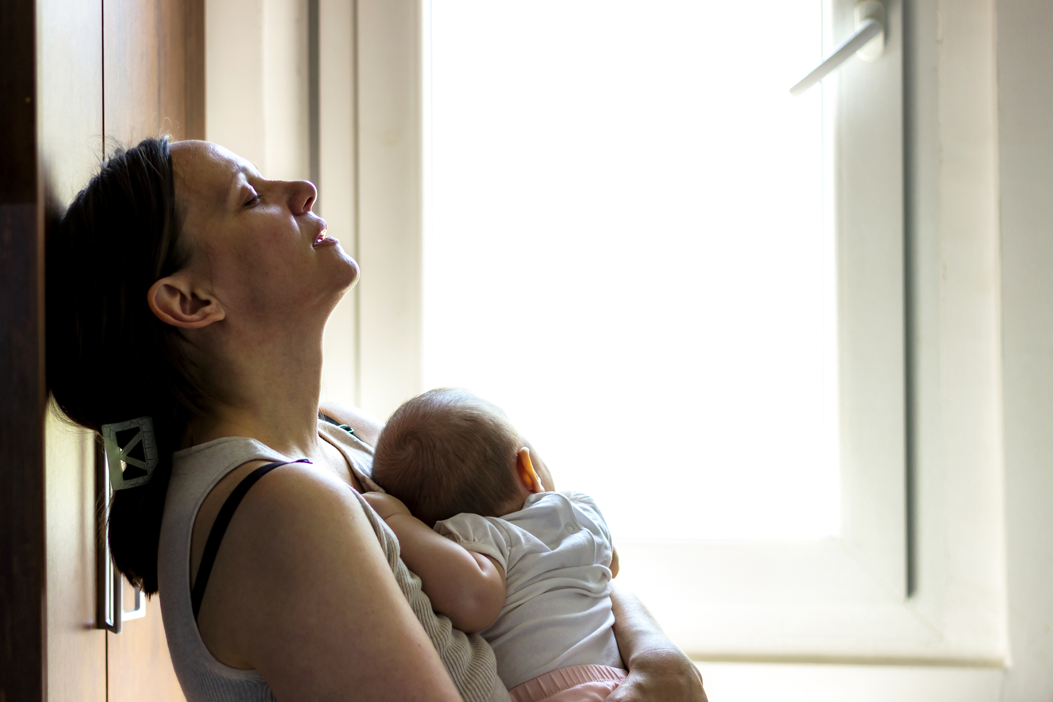 Tired concerned mother rocking sleeping baby. A mother sitting by a window, leaning back with her eyes closed in an expression of exhaustion, while holding her baby close to her chest. The soft natural light coming through the window highlights the quiet and intimate moment of caregiving and fatigue.