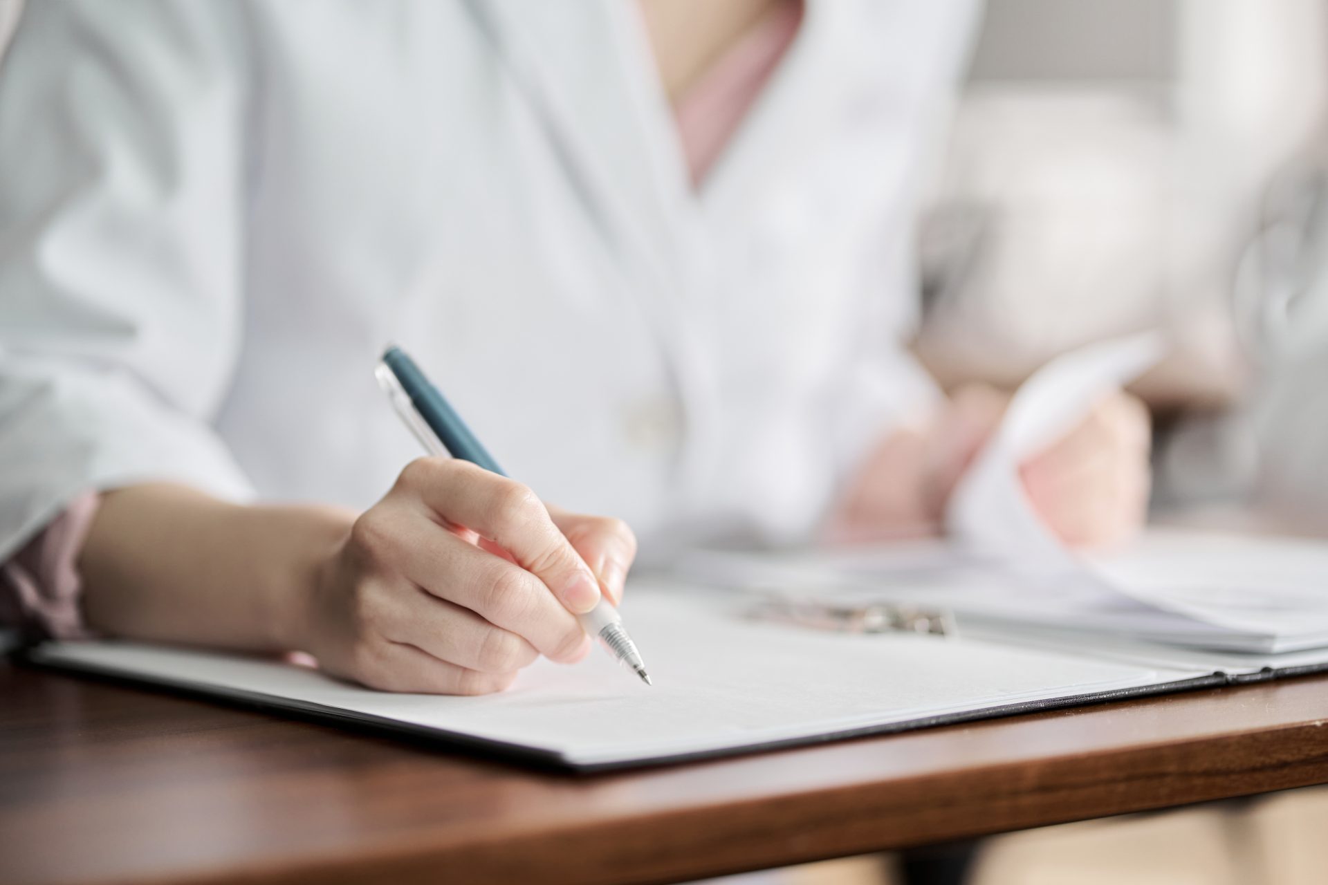iStock-1353348897 Close-up of a healthcare professional's hand holding a pen while writing on a clipboard. The individual is wearing a white coat, with documents and a blurred background indicating a clinical or office setting.