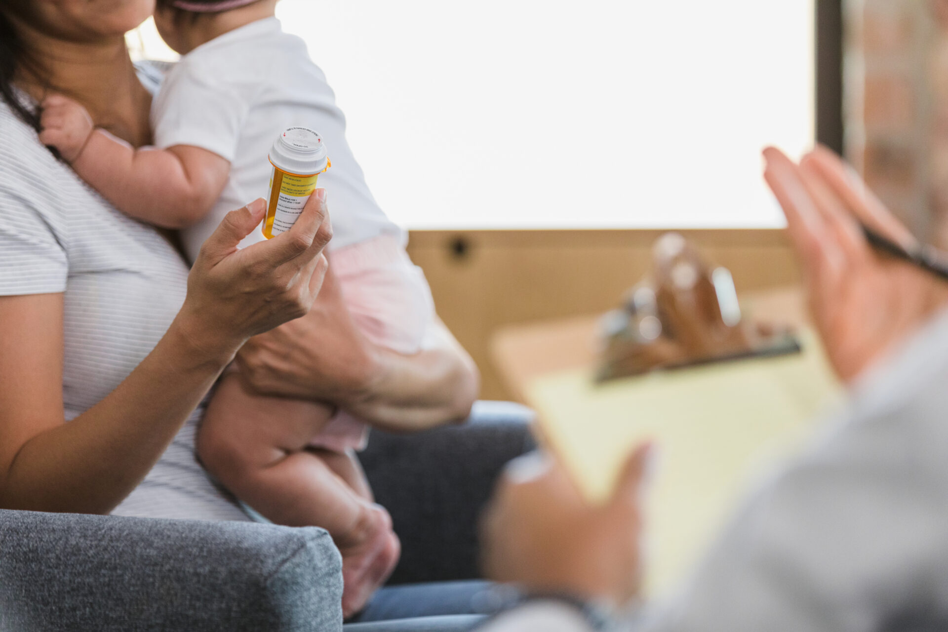 Unrecognizable woman holds prescription bottle while listening to unrecognizable psychiatrist A mother holding her baby while sitting in a chair, discussing a prescription bottle with a healthcare professional. The healthcare provider gestures while holding a clipboard, emphasizing communication about medication and postpartum care in a supportive setting.