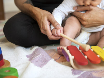 Parent Bonding With Infant During Early Childhood Development Activity—helping Baby Play Colorful Wooden Xylophone For Sensory And Motor Skills Growth. Montessori-inspired Musical Toy On Soft Play Mat With Educational Stacking Rings Nearby.