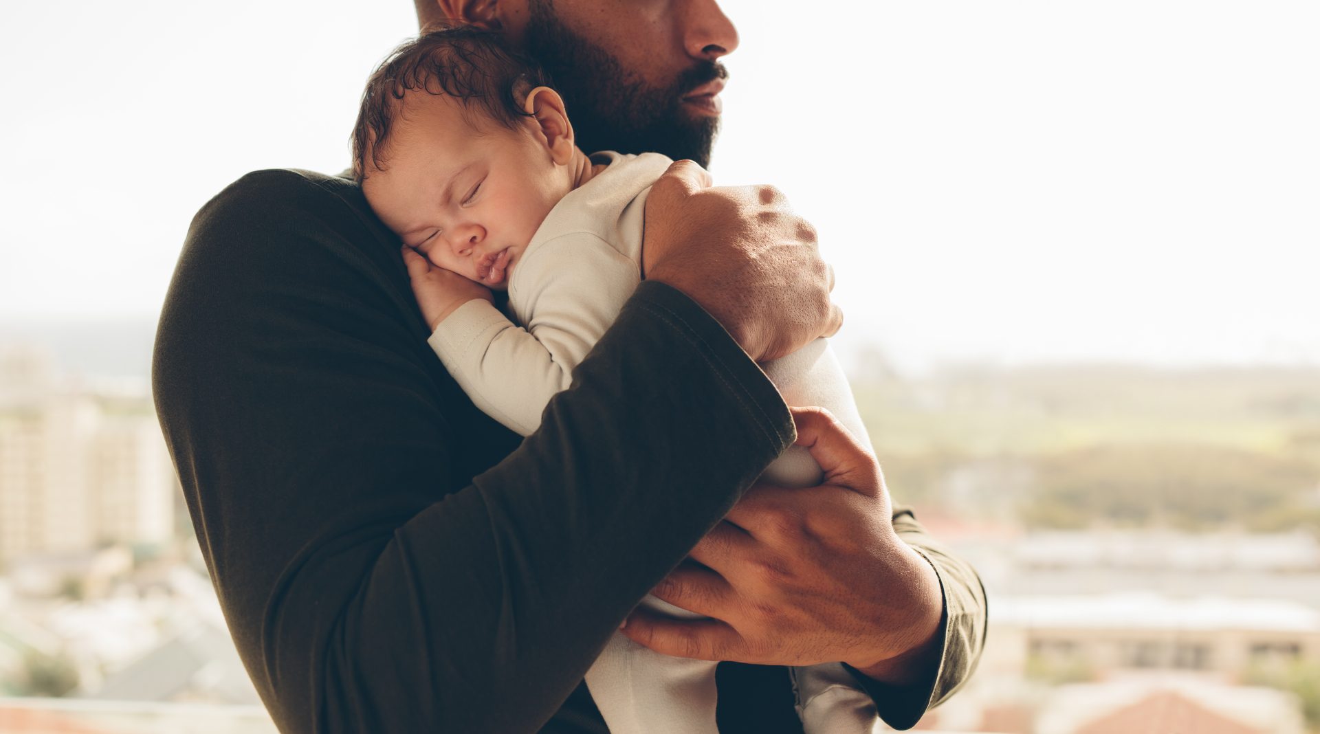 iStock-882871236 A father tenderly holding his sleeping baby close to his chest, creating a sense of warmth and security. The baby rests peacefully in a white onesie, while the father wears a dark shirt. The background shows a blurred cityscape, emphasizing a calm and nurturing moment.