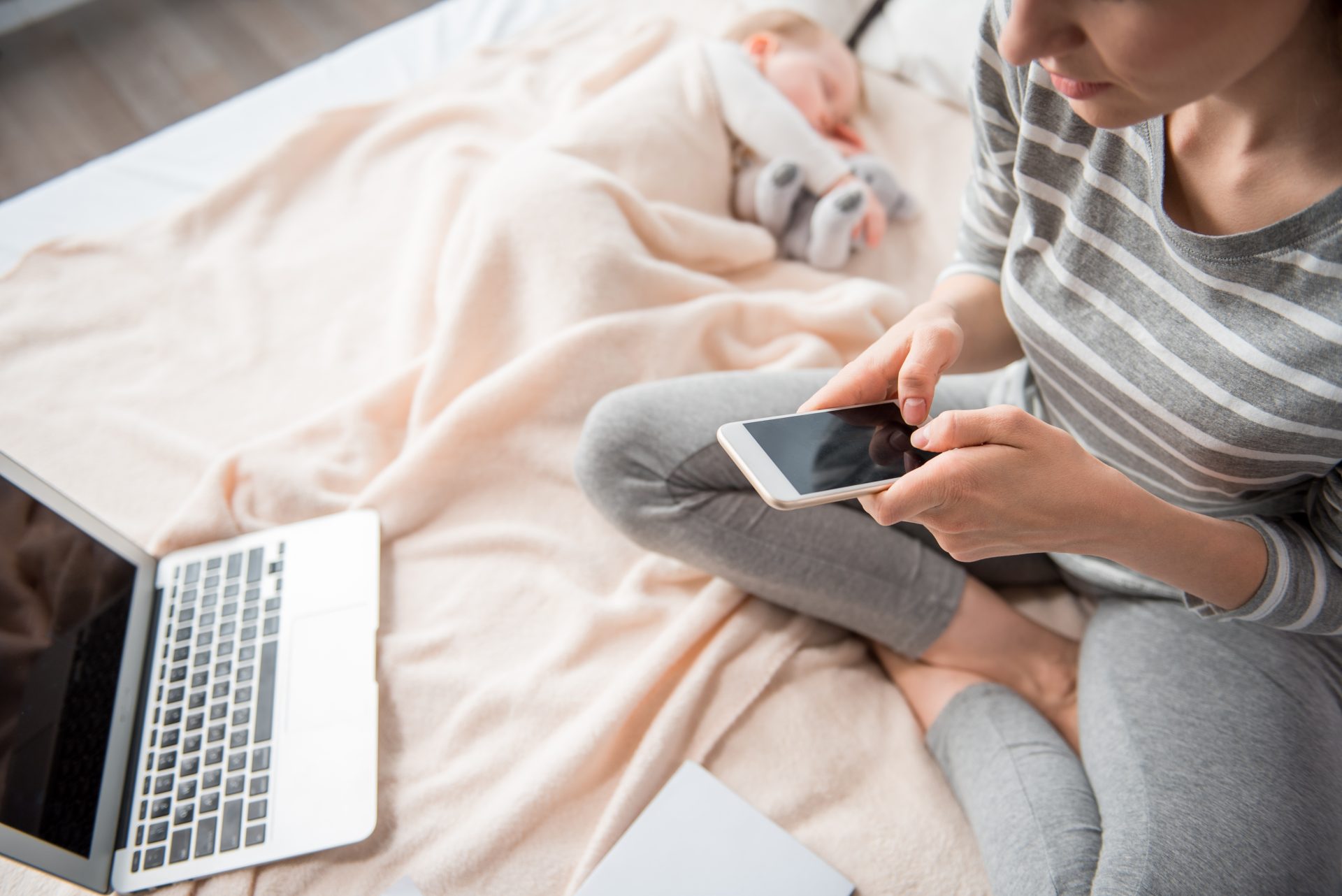 A mother in a striped shirt and gray leggings sitting on a bed, using her smartphone while a laptop rests nearby. In the background, her baby sleeps peacefully under a soft beige blanket, creating a serene and multitasking moment.