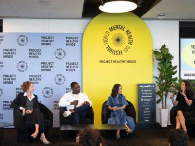 A Panel Discussion At The World Mental Health Day Festival, Hosted By Project Healthy Minds. Four Speakers Are Seated On Stage: A Woman In A Business Suit, A Man In Casual Attire, A Woman In A Denim Dress, And Another Woman In A Black Dress. The Vibrant Backdrop Includes The 'Project Healthy Minds' Logo And A Yellow Arch With The 'World Mental Health Day Festival' Branding. The Session Addresses Topics Related To Mental Health And Parenting.