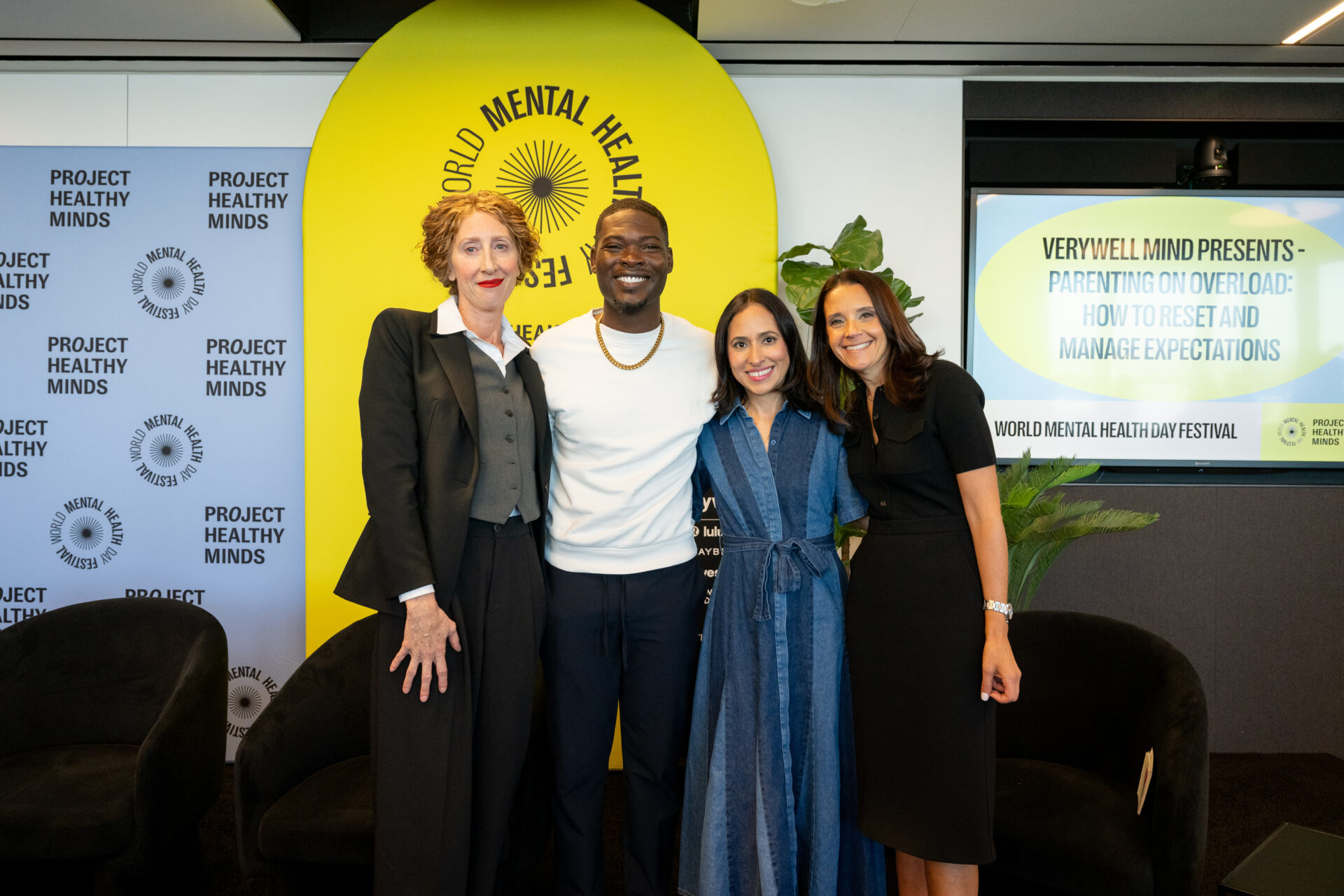 A group photo featuring four individuals standing together at the World Mental Health Day Festival, hosted by Project Healthy Minds. The backdrop includes event branding and a presentation screen with the topic 'Parenting on Overload: How to Reset and Manage Expectations,' highlighting the theme of mental health awareness.