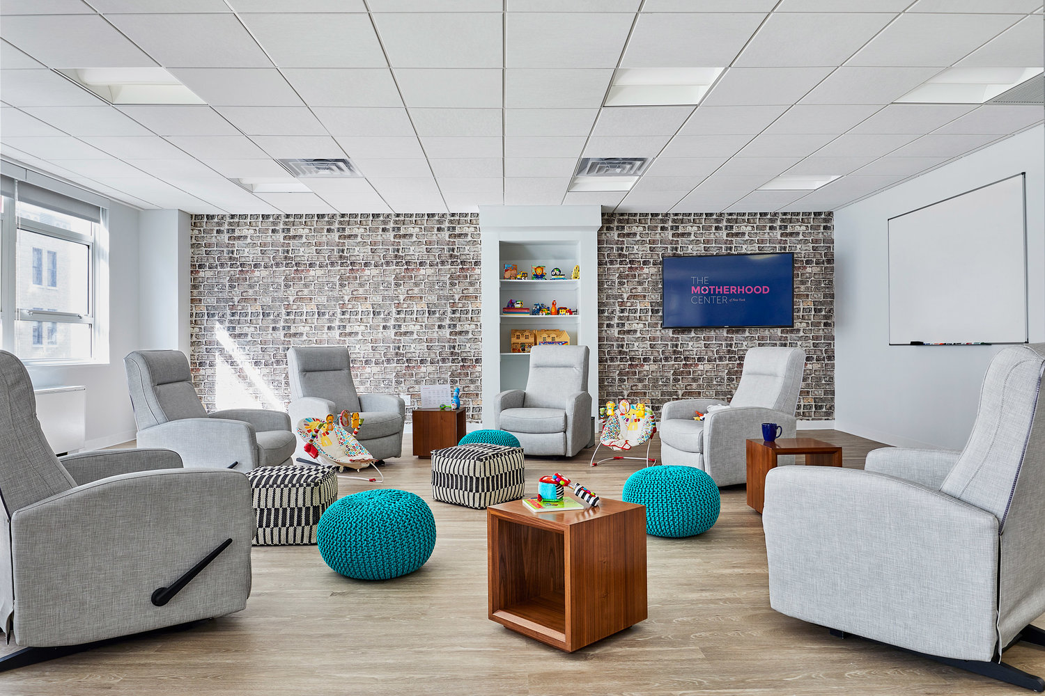The_Motherhood_Center_Room A modern and inviting therapy room at The Motherhood Center's Day Program group room. The space features comfortable gray recliners arranged in a circle, colorful knitted poufs, and wooden side tables. A brick accent wall includes shelves with toys and a screen displaying 'The Motherhood Center' logo. The room is brightly lit with natural light from large windows, creating a warm and welcoming environment for group therapy sessions and support meetings.