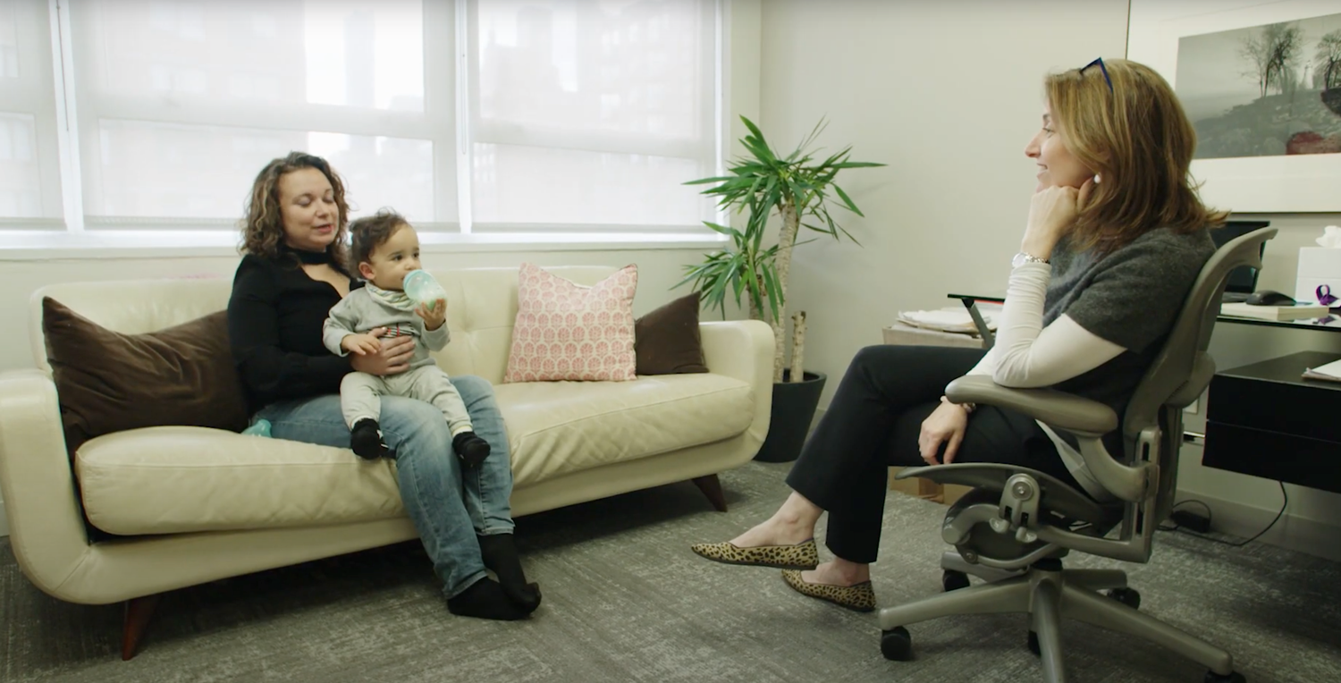 DayProgram_IndividualTherapy A serene therapy session in progress. On the left, a mother sits on a cream-colored couch holding her baby, who is drinking from a bottle. She is engaged in conversation with the therapist seated on the right in a comfortable chair, listening attentively. The room is warmly lit, with a large window providing natural light. Decorative accents include a patterned pillow, a potted plant, and a minimalist desk setup in the background, creating a welcoming and supportive environment.