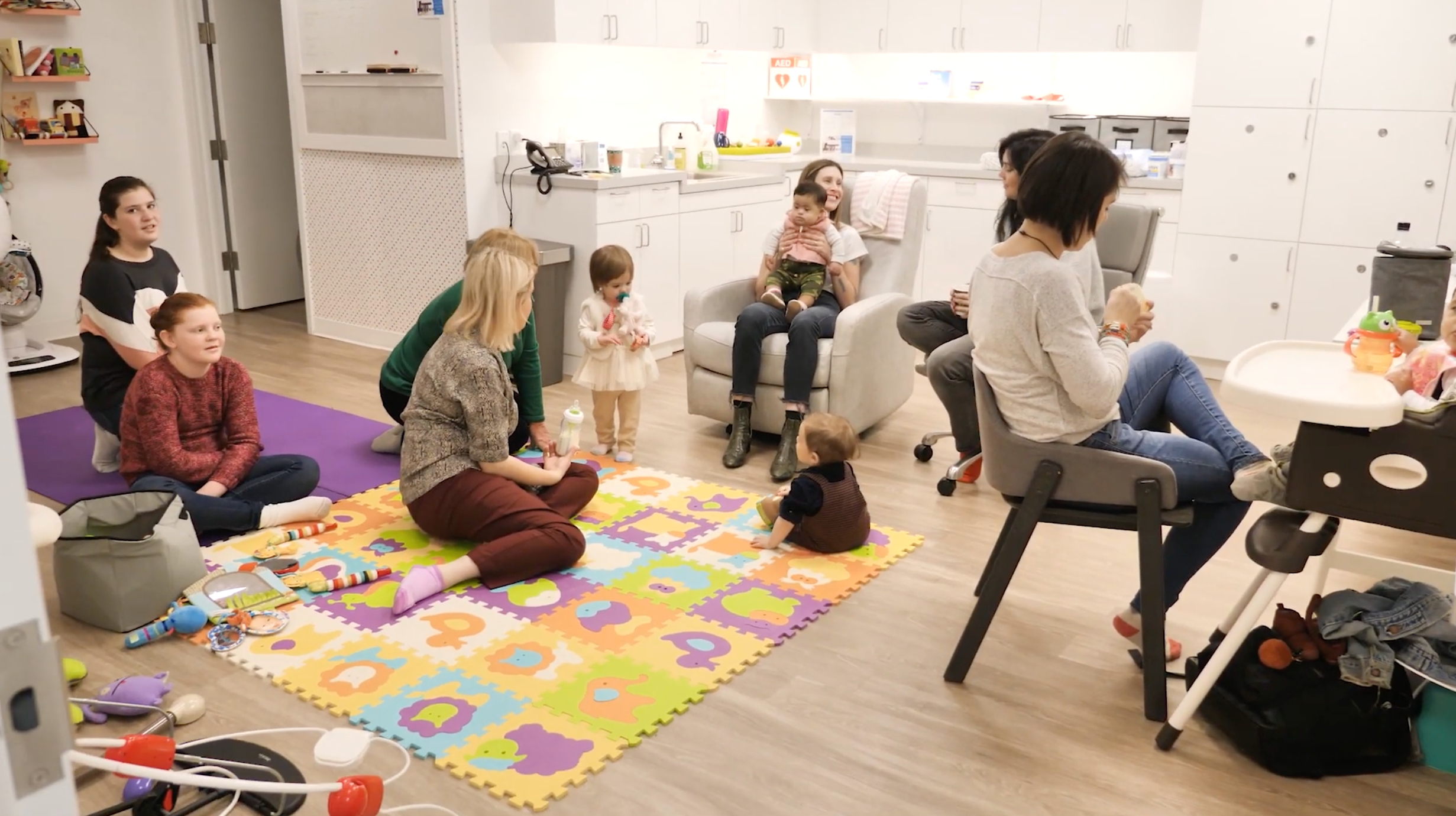 FromVideo_GroupShot A group of moms and babies gathered in The Motherhood Center's nursery. The room features a colorful play mat with toys, comfortable seating, and a bright, organized space with white cabinets and countertops. Some mothers are sitting on the floor interacting with their children, while others are seated, holding their babies or chatting. The scene emphasizes community, support, and care in a welcoming environment.
