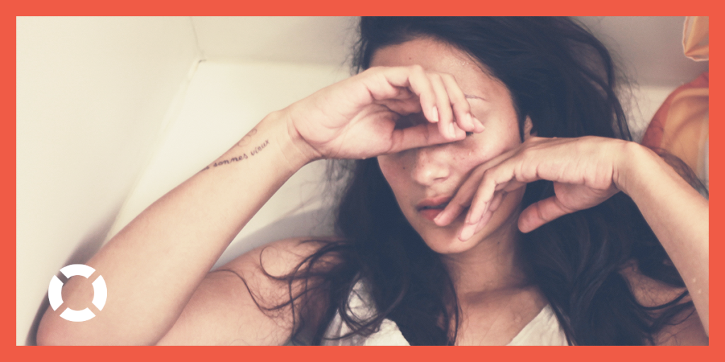 A close up photograph of a woman laying in a tub, with her hands raised to cover her face